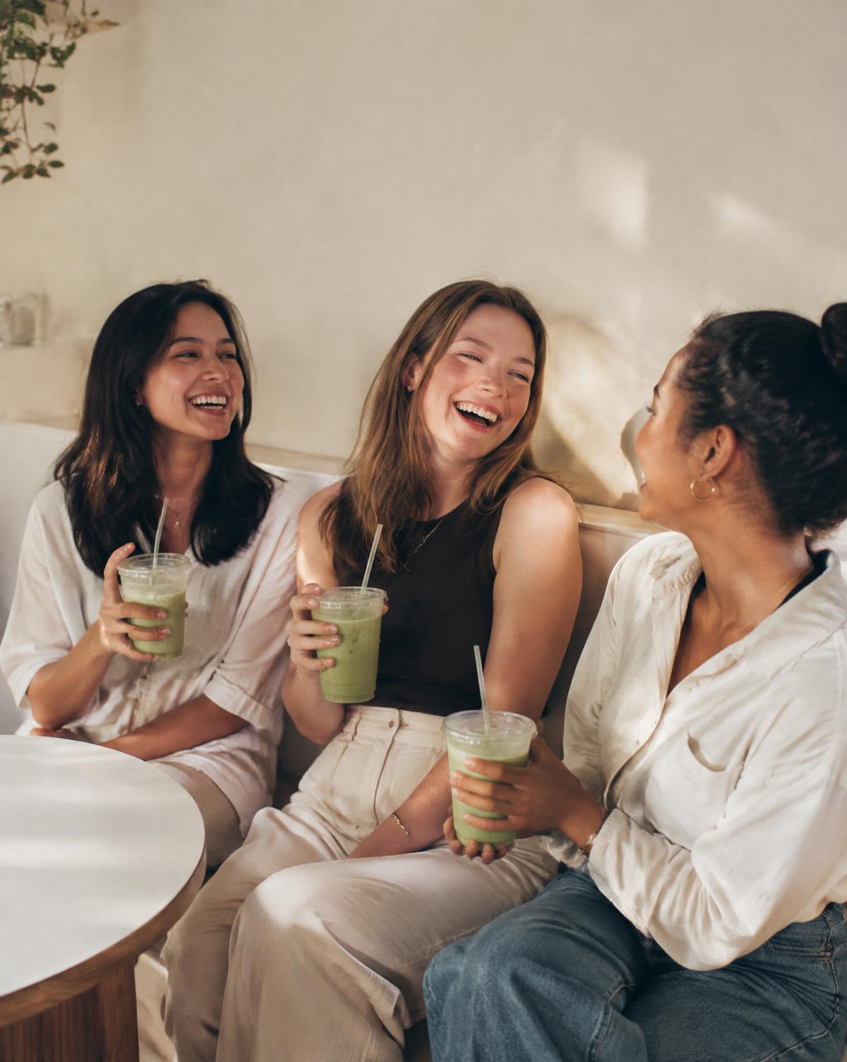 Three friends laughing together with iced matcha drinks in a bright café