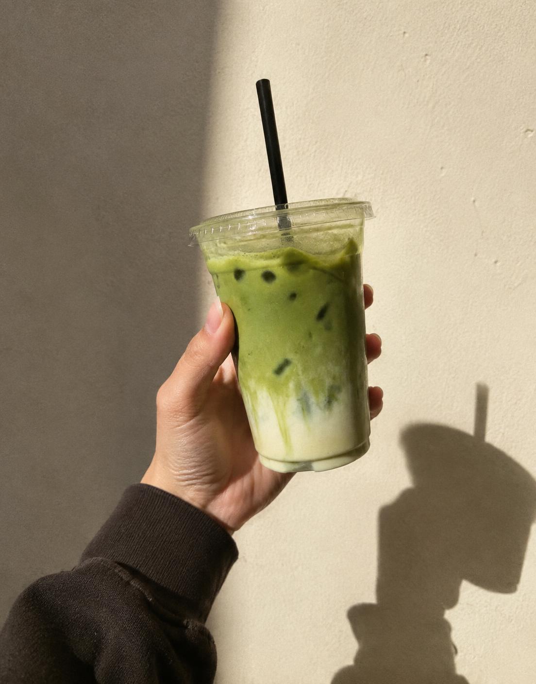 Hand holding an iced matcha to-go cup against a sunlit wall