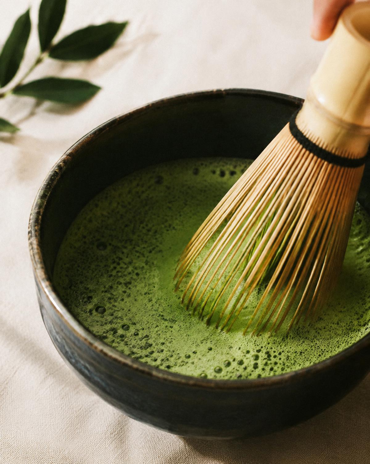 Bamboo whisk frothing bright green matcha in a dark ceramic bowl
